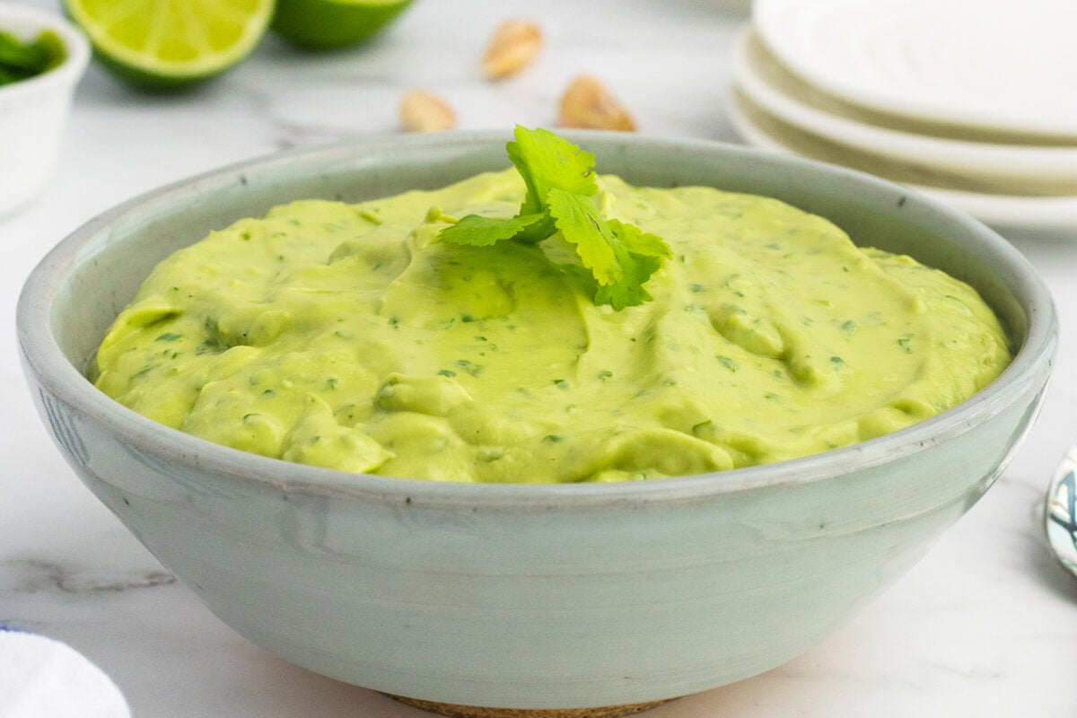 A light green bowl filled with creamy guacamole, garnished with fresh cilantro, sits on a white surface with limes, pistachios, and stacked white plates in the background.