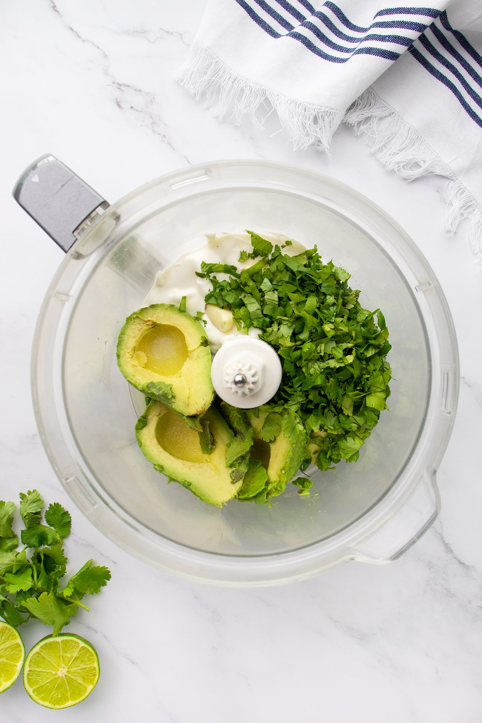 A food processor bowl containing halved avocados, chopped cilantro, and a dollop of mayonnaise, on a white marble surface with a striped towel, fresh cilantro, and sliced lime nearby.