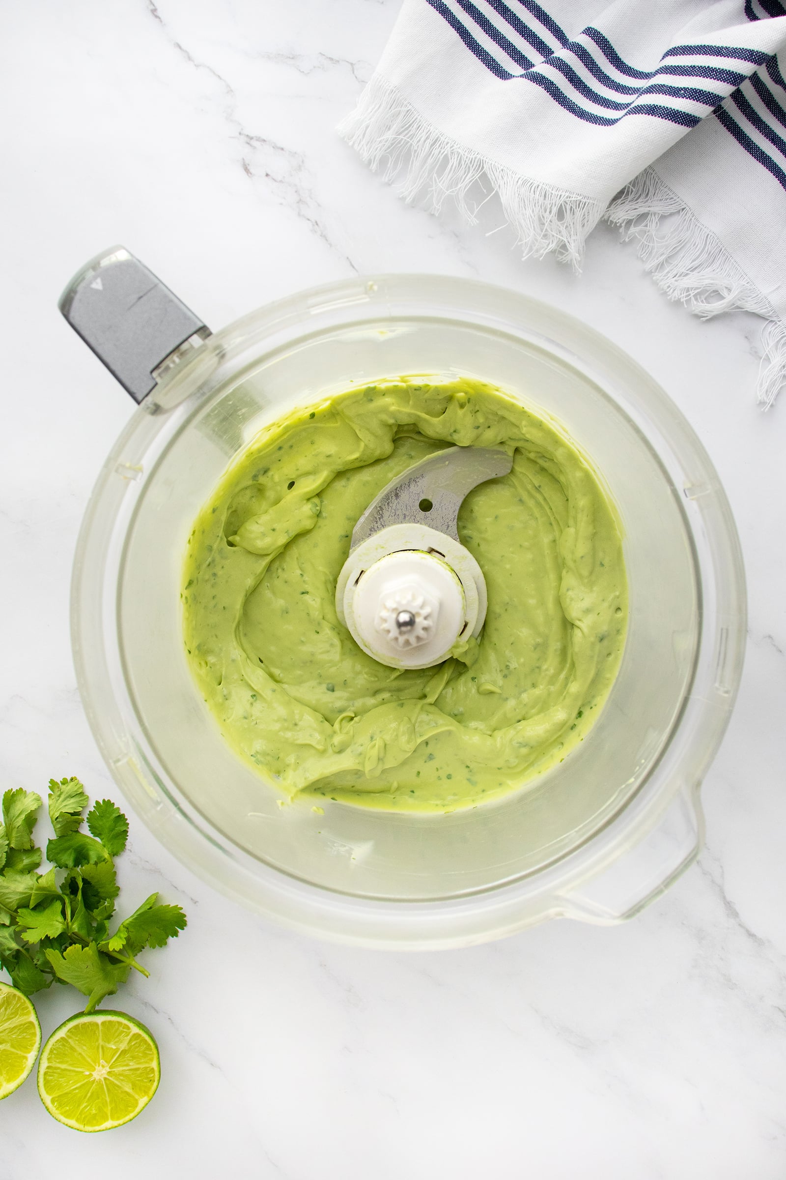 A glass food processor bowl filled with creamy green avocado sauce, surrounded by fresh cilantro, lime halves, and a striped white kitchen towel on a marble countertop.