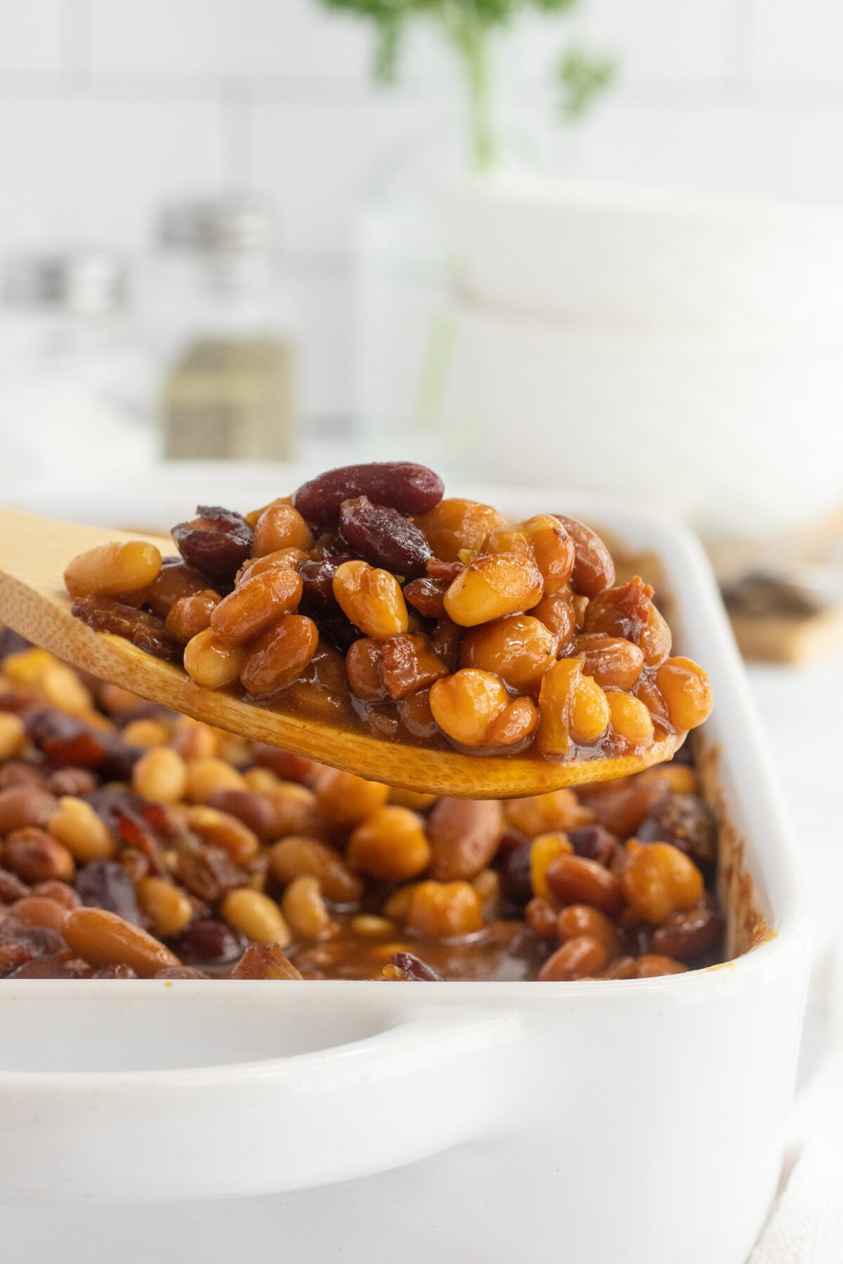 A wooden spoon lifts a serving of baked beans from a white ceramic baking dish filled with mixed beans in sauce. Blurred dishes and condiments are visible in the background.