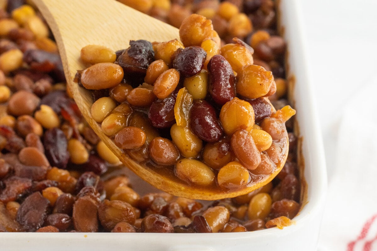 A wooden spoon scooping a mix of baked beans in sauce from a white dish, with various types of beans visible, including kidney beans and navy beans.