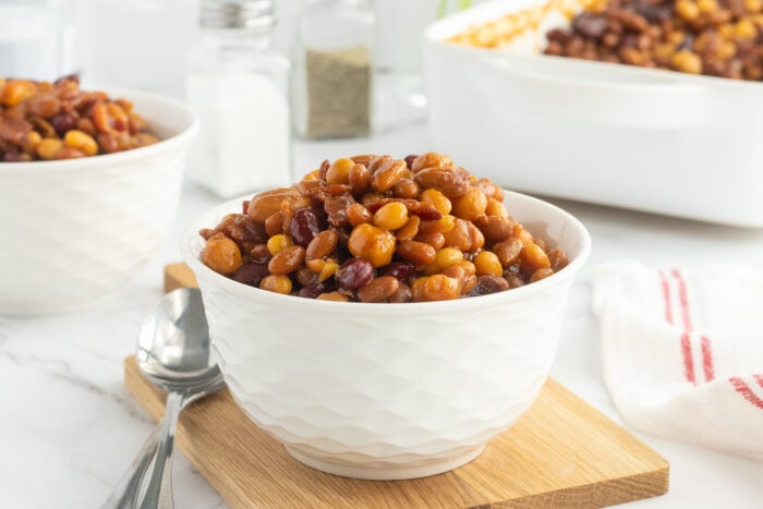 A white bowl filled with baked beans sits on a wooden board, with two spoons beside it. In the background, theres another bowl of beans, salt and pepper shakers, and a white dish.