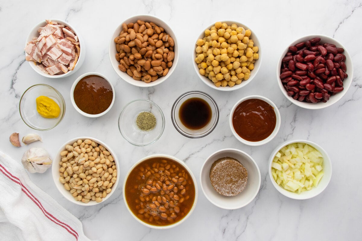 An assortment of ingredients in bowls on a white surface, including beans, chickpeas, kidney beans, bacon, chopped onion, garlic, sauces, mustard, seasoning, and spices, arranged in a grid pattern.