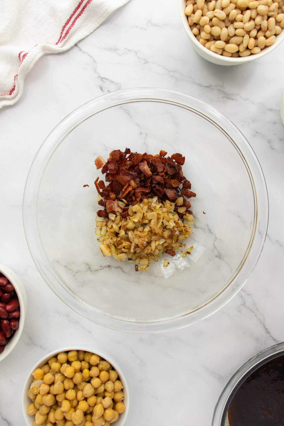 A clear glass bowl contains cooked chopped bacon and sautéed onions. Surrounding the bowl are smaller bowls filled with navy beans, kidney beans, chickpeas, and a dark sauce, all on a white marble surface.