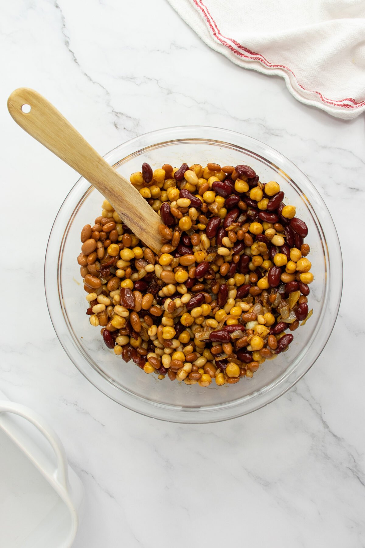A glass bowl filled with a mixed bean salad and a wooden spoon, set on a white marble surface with a white cloth featuring a red trim in the corner.