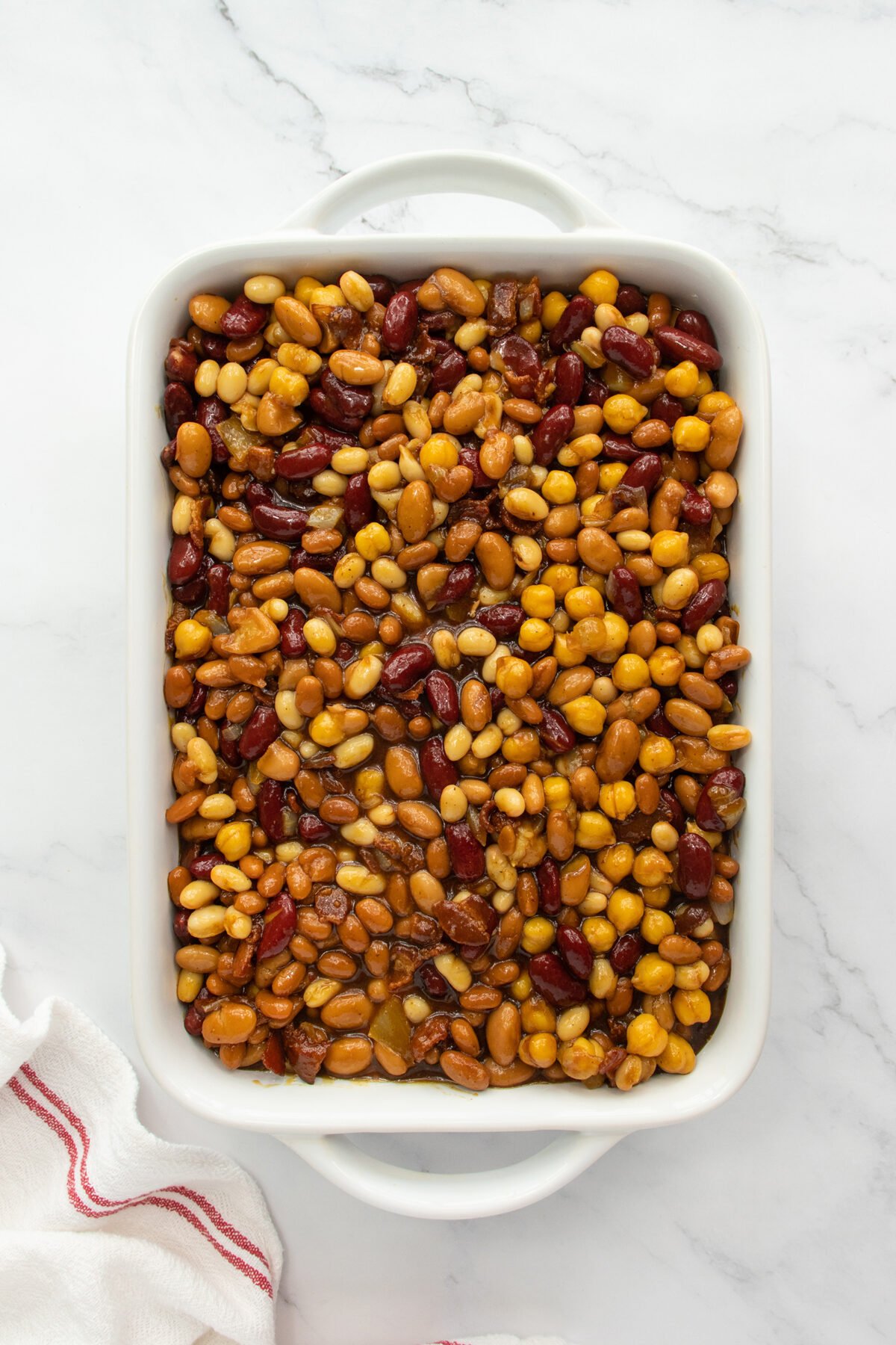 A white rectangular baking dish filled with a mixture of baked beans in various shades of brown, red, and yellow, placed on a white marble surface with a striped cloth nearby.