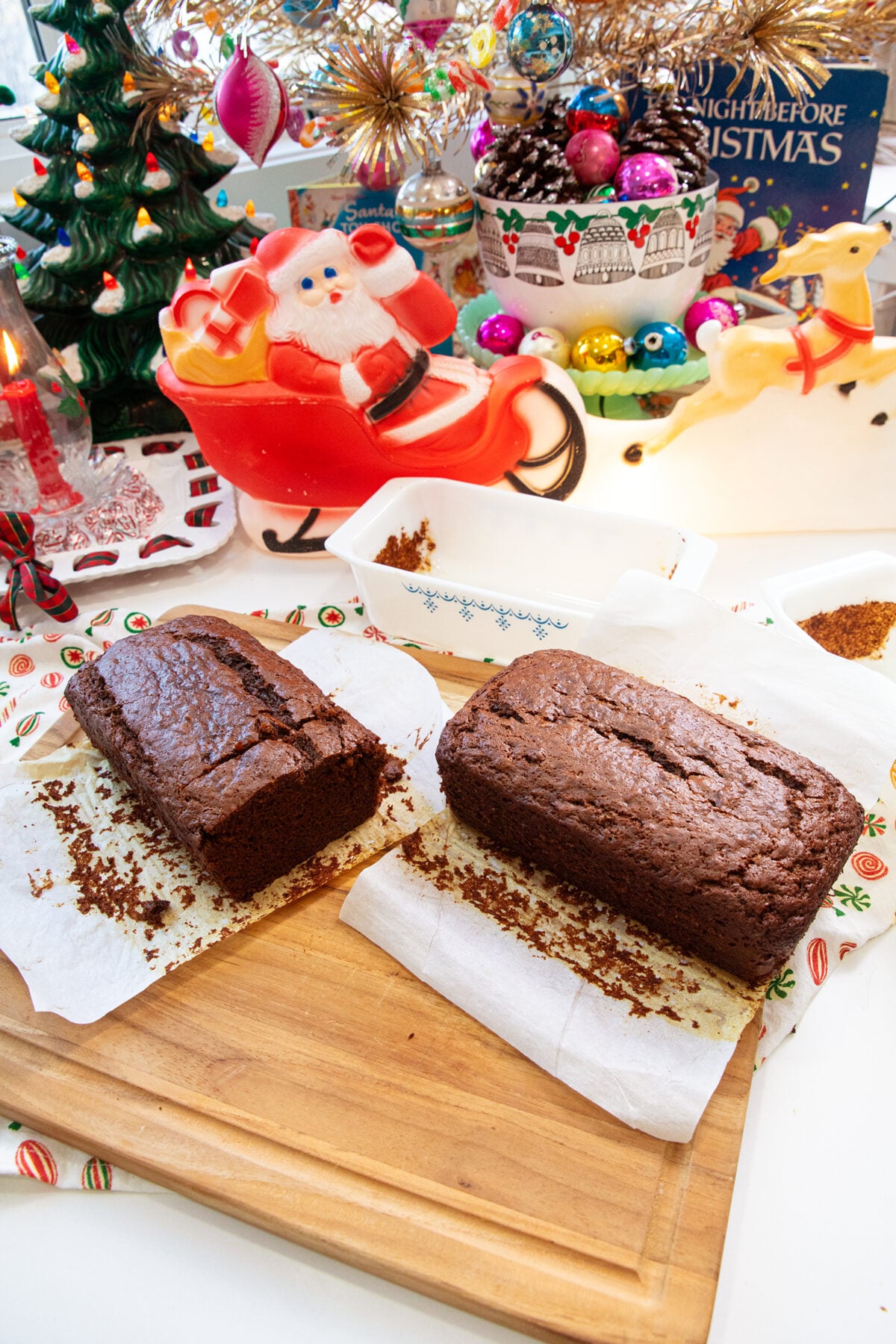 Two chocolate loaves on parchment paper sit on a wooden board, surrounded by festive Christmas decorations, including a Santa figurine in a sleigh, a Christmas tree, ornaments, and a reindeer figure.