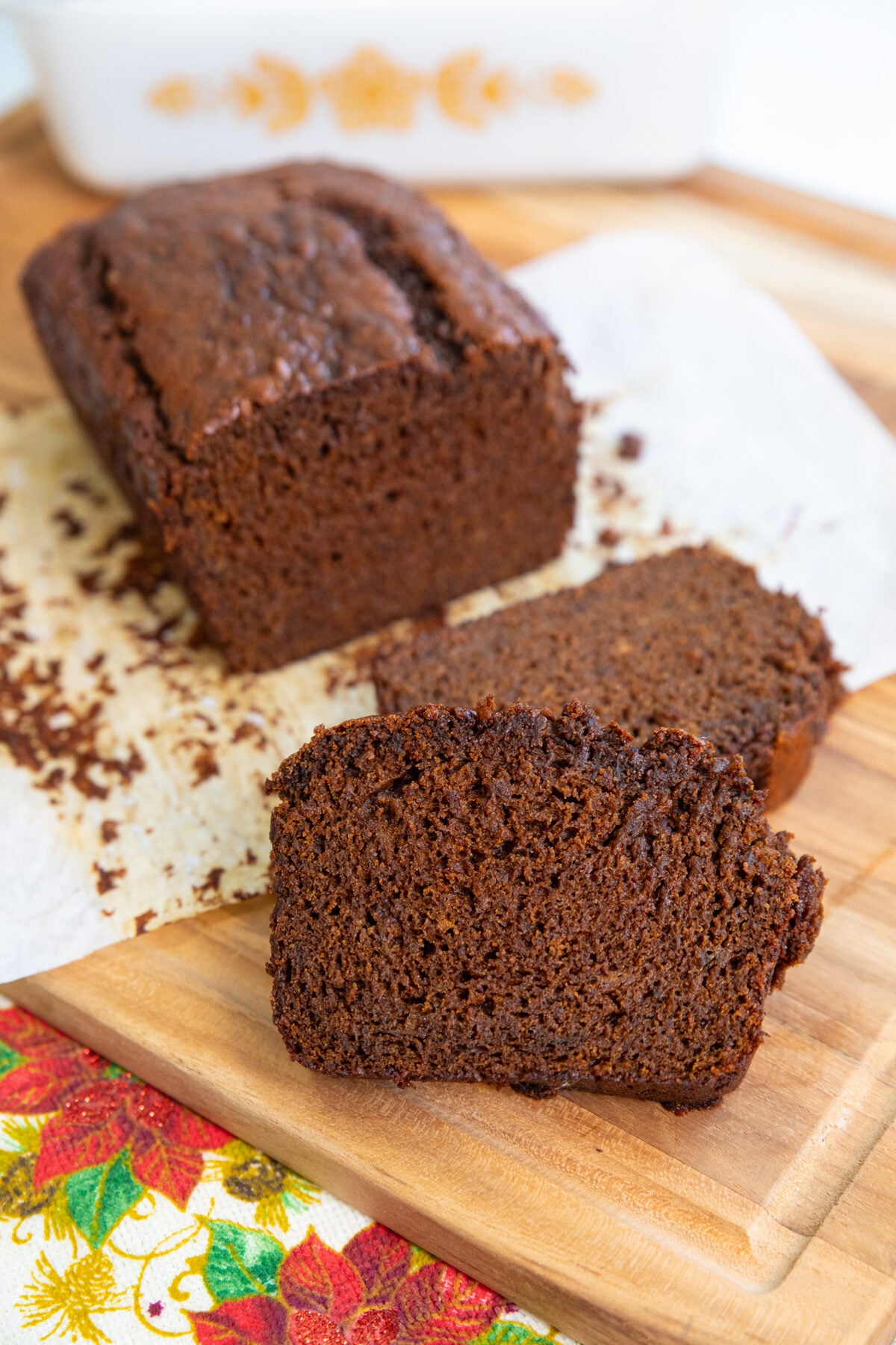 A loaf of chocolate banana bread sits on parchment paper atop a wooden cutting board, with two slices cut and placed in front. A colorful floral napkin is partially visible in the corner.