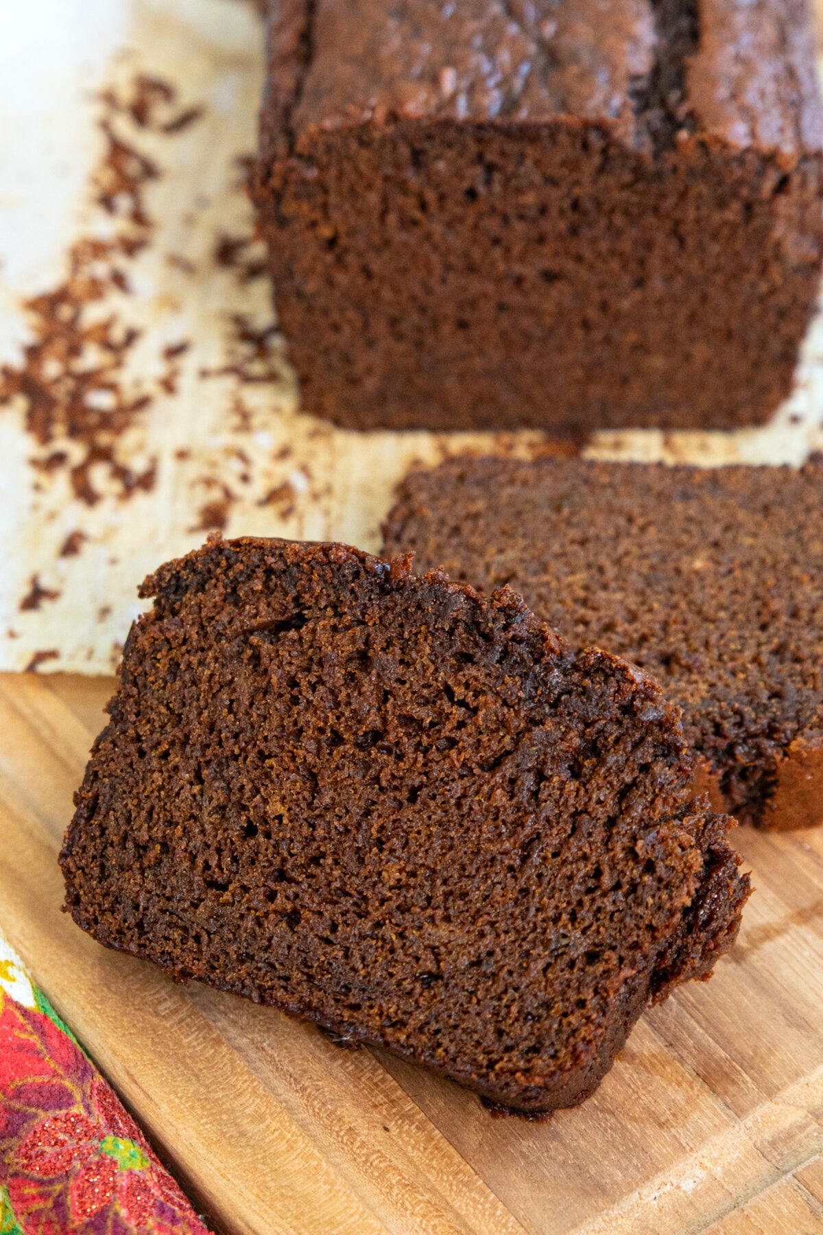 A close-up of sliced chocolate banana bread on a wooden cutting board, with the rest of the loaf and parchment paper in the background. The bread looks moist and rich in color.