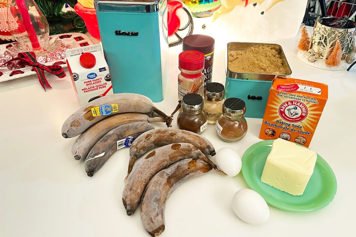 A selection of baking ingredients on a counter: frozen bananas, eggs, butter, baking soda, spices, brown sugar, flour, cocoa powder, baking powder, heavy cream, and kitchen containers.