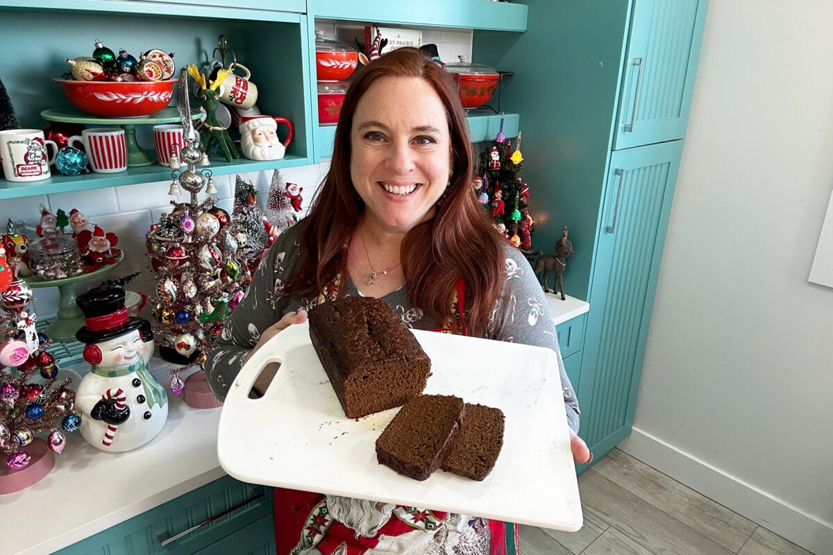 A smiling woman with long red hair holds a white cutting board with a sliced loaf of bread in a festive kitchen decorated with Christmas ornaments and holiday-themed items.