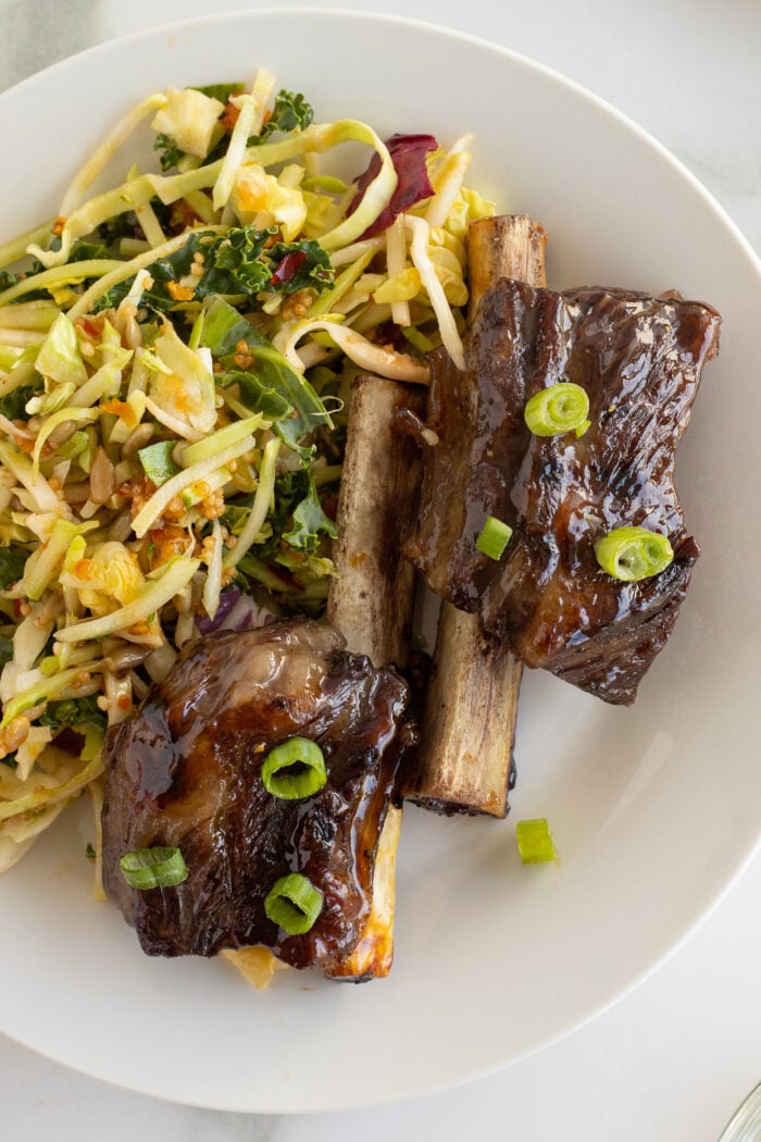 A white plate with two glazed beef short ribs garnished with chopped green onions, served alongside a fresh vegetable slaw with greens and shredded vegetables.