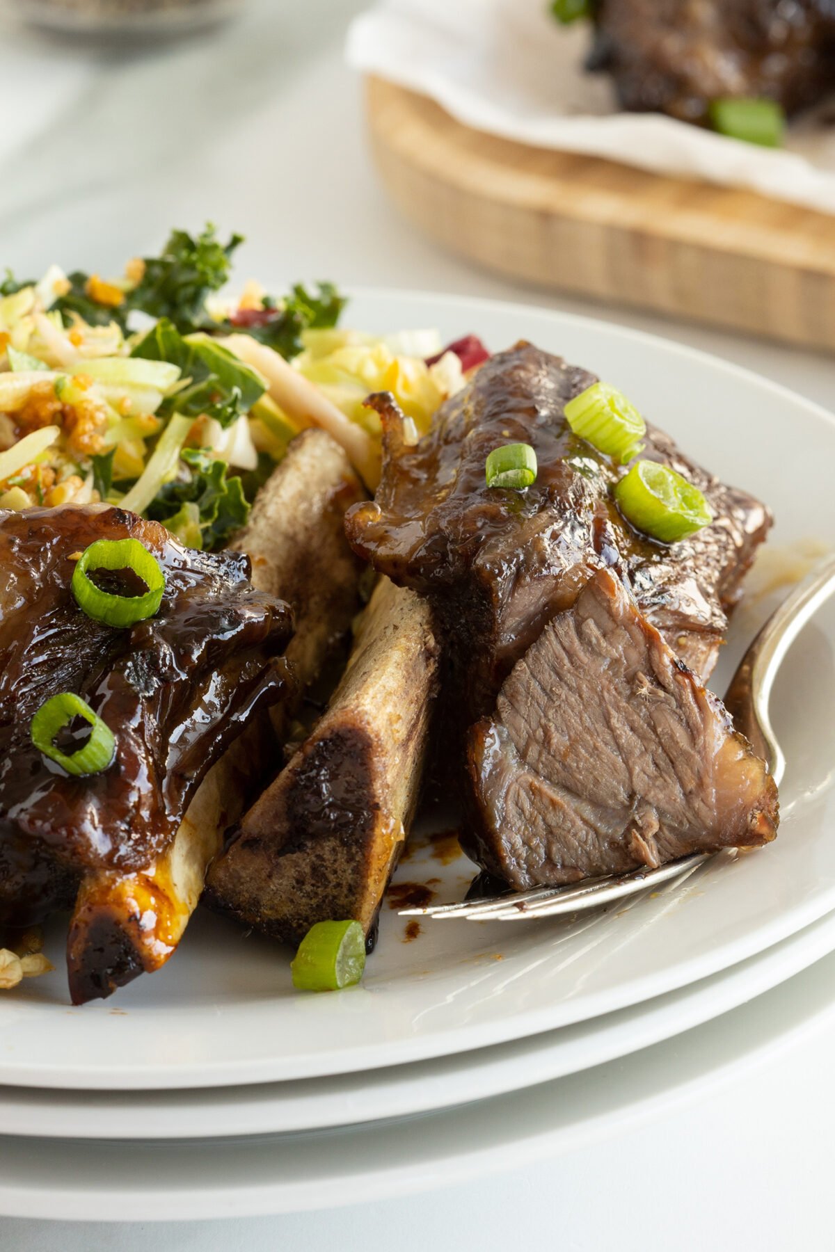 A plate of glazed beef short ribs garnished with chopped green onions, served with a fresh salad featuring kale and apple slices. A fork rests beside the ribs on the white plate.