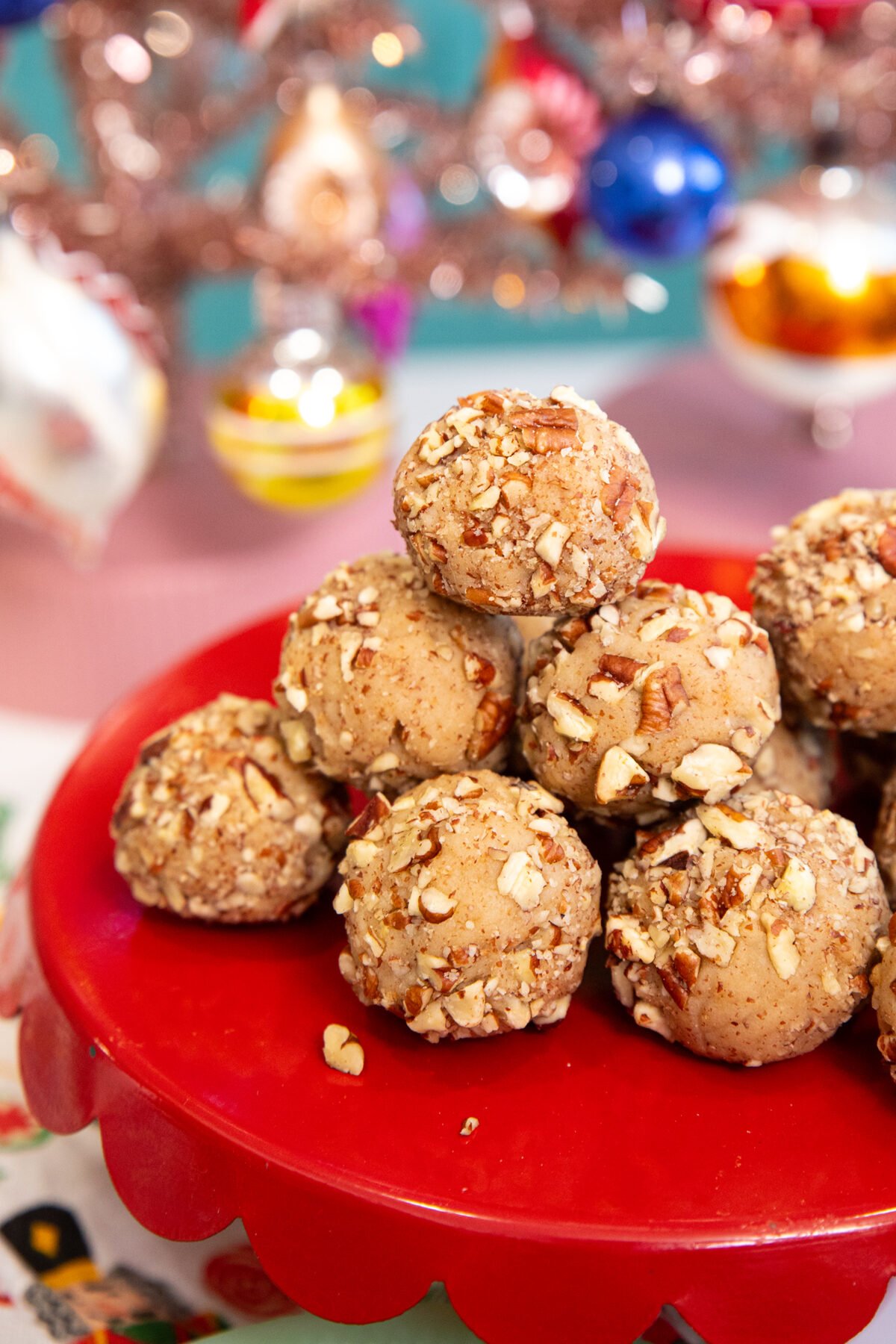 A stack of round, nut-coated cookies arranged on a red cake stand with colorful, blurred holiday ornaments in the background.
