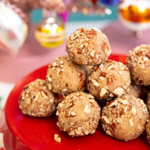 A stack of round, nut-coated cookies arranged on a red cake stand with colorful, blurred holiday ornaments in the background.