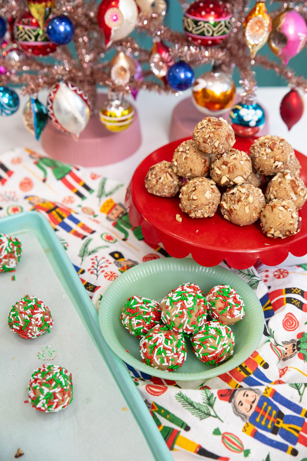 A festive holiday scene with colorful Christmas ornaments, two plates of round cookies—one with red, white, and green sprinkles, the other with chopped nuts—on a decorative tablecloth with holiday patterns.
