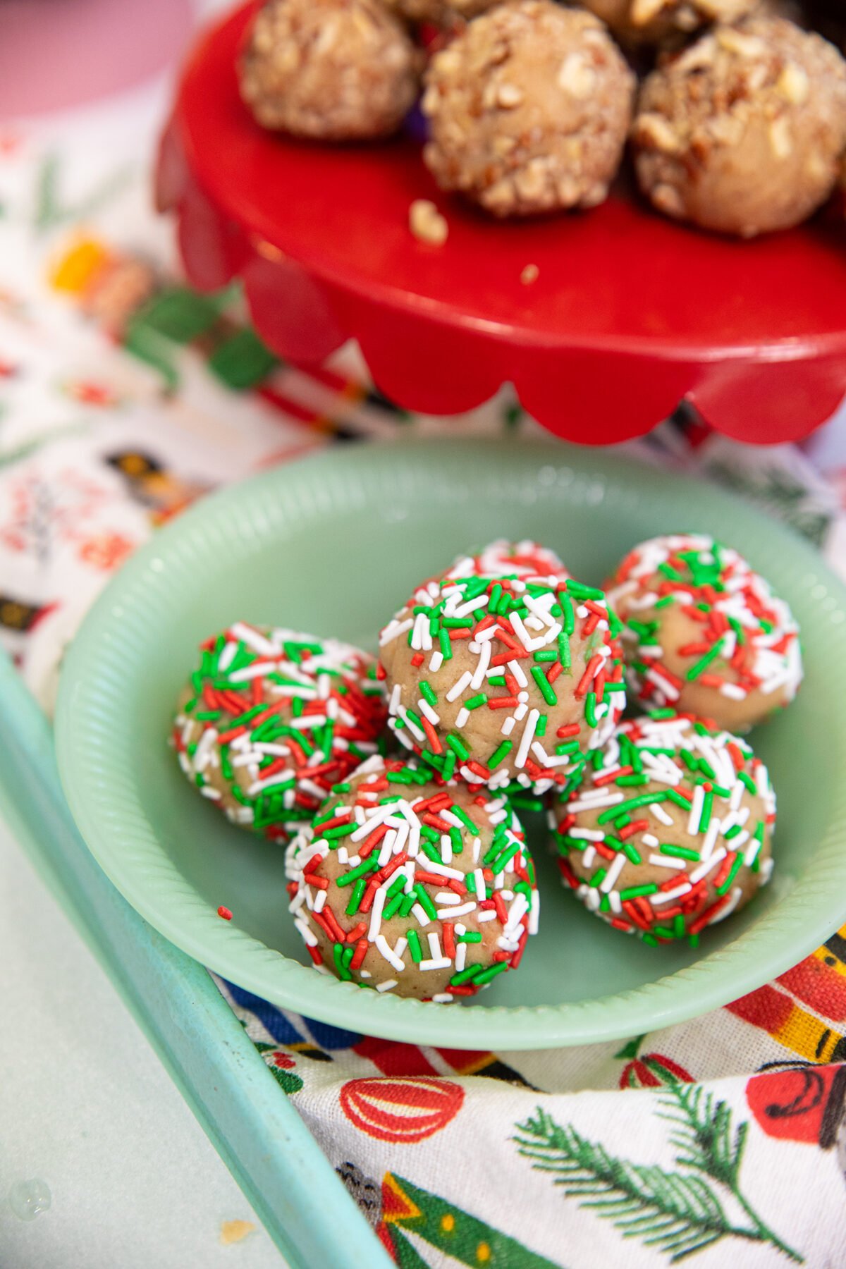 A green plate holds round holiday treats covered in red, green, and white sprinkles. In the background, more treats sit on a red stand, and a festive tablecloth with holiday designs is visible.