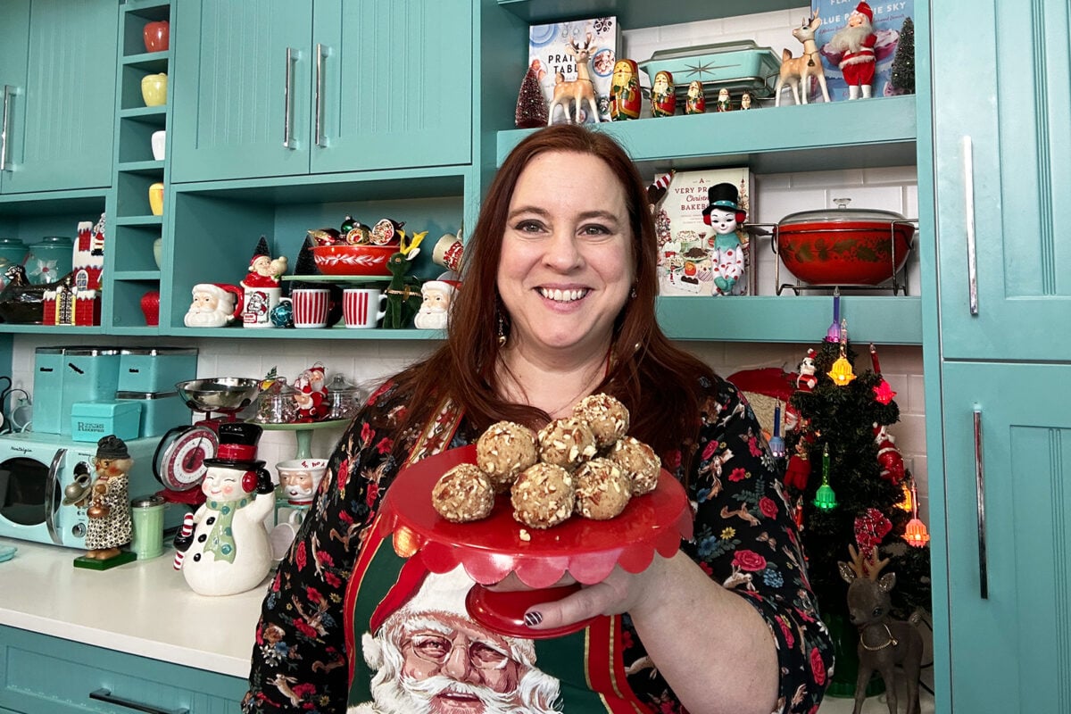 A smiling woman in a festive kitchen holds a red tray of homemade treats. She wears a Santa-themed apron, and holiday decorations, including Santa figurines, are displayed on turquoise cabinets behind her.