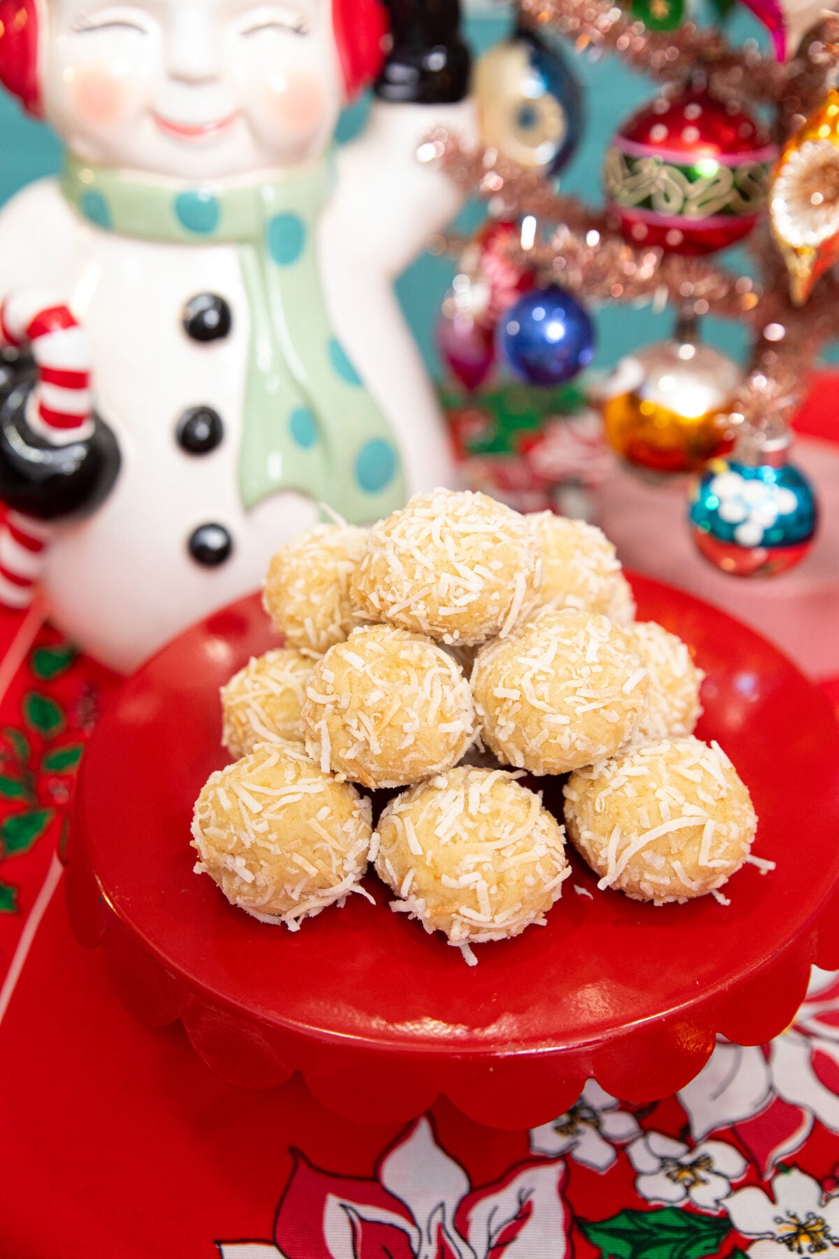A red plate holds coconut-covered dessert balls. In the background, there is a cheerful snowman figurine with earmuffs and a small, decorated Christmas tree with colorful ornaments.