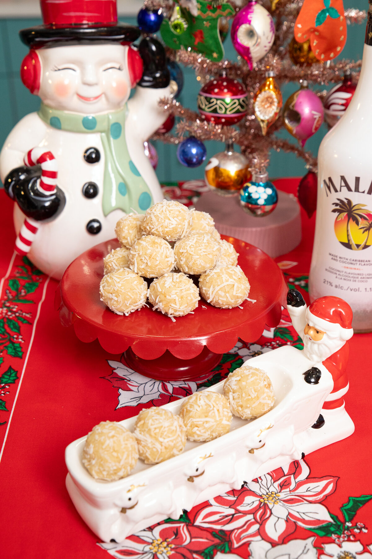 A festive holiday table displays coconut-covered treats on red platters, surrounded by a snowman cookie jar, a small decorated Christmas tree, and a bottle of Malibu. The tablecloth features poinsettia designs.