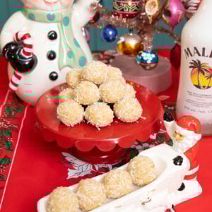 A festive holiday table displays coconut-covered treats on red platters, surrounded by a snowman cookie jar, a small decorated Christmas tree, and a bottle of Malibu. The tablecloth features poinsettia designs.