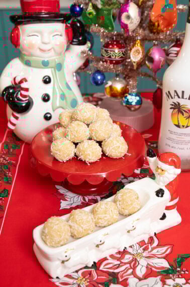 A festive holiday table displays coconut-covered treats on red platters, surrounded by a snowman cookie jar, a small decorated Christmas tree, and a bottle of Malibu. The tablecloth features poinsettia designs.