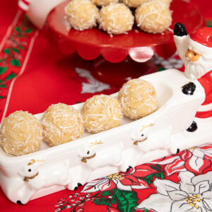 A festive ceramic dish shaped like Santa’s sleigh, filled with four coconut-covered dessert balls, sits on a red holiday tablecloth. More treats are on a red pedestal dish in the background.