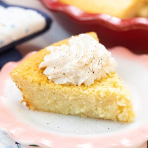 A slice of cornbread topped with whipped cream sits on a white plate with a pink rim. A larger dish of cornbread is seen in the background. The scene is decorated with a festive cloth.
