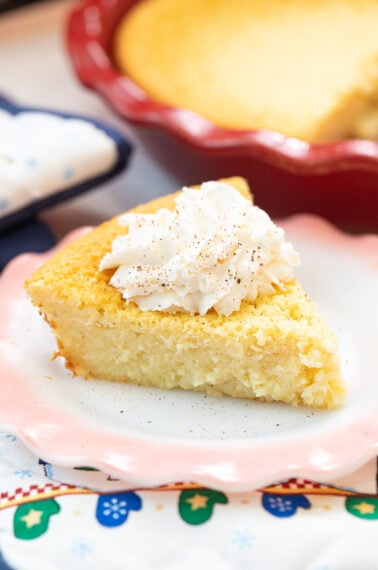 A slice of cornbread topped with whipped cream sits on a white plate with a pink rim. A larger dish of cornbread is seen in the background. The scene is decorated with a festive cloth.