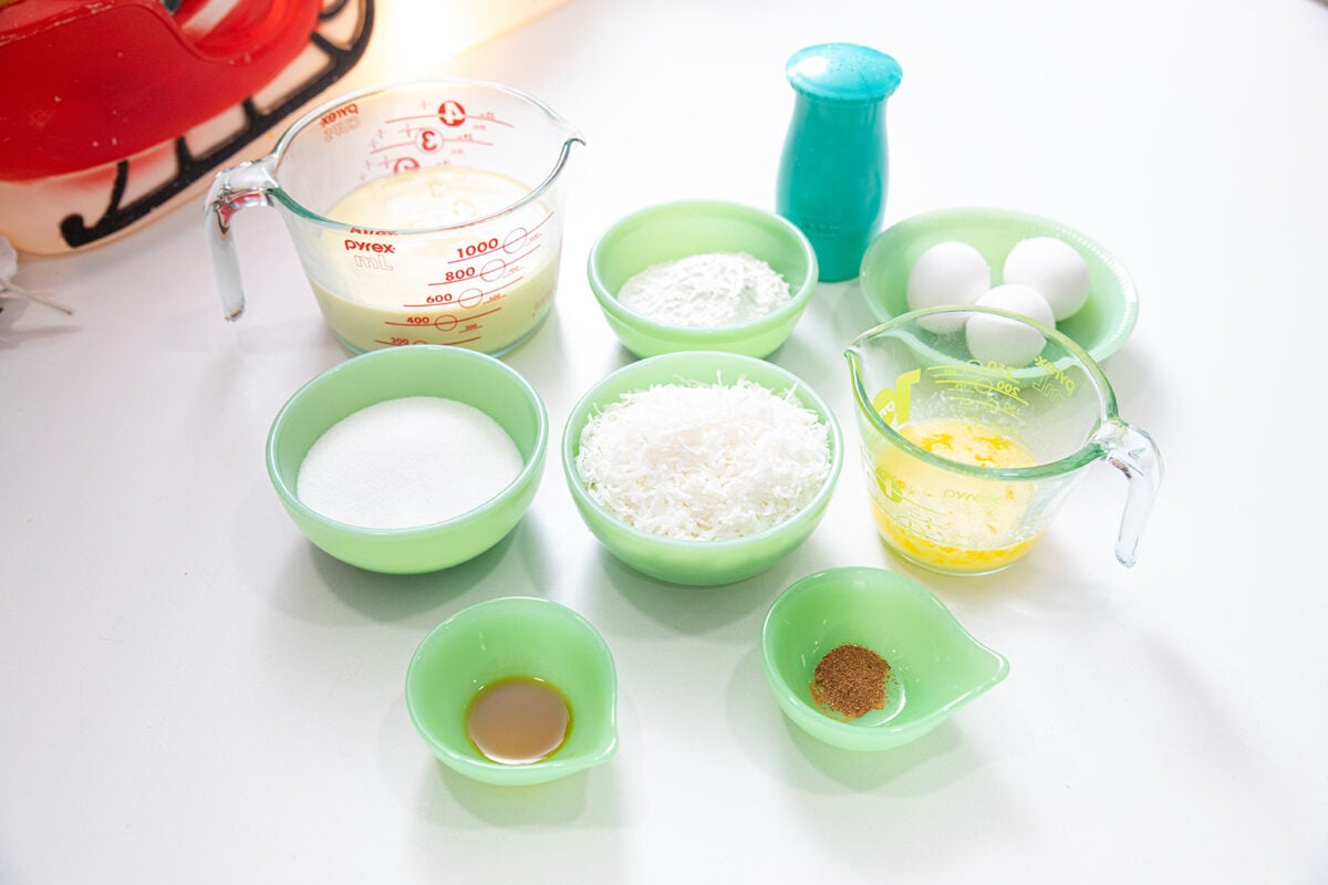 A collection of baking ingredients in green bowls and measuring cups, including milk, flour, eggs, sugar, shredded coconut, melted butter, vanilla extract, spices, and a blue salt shaker, arranged on a white countertop.