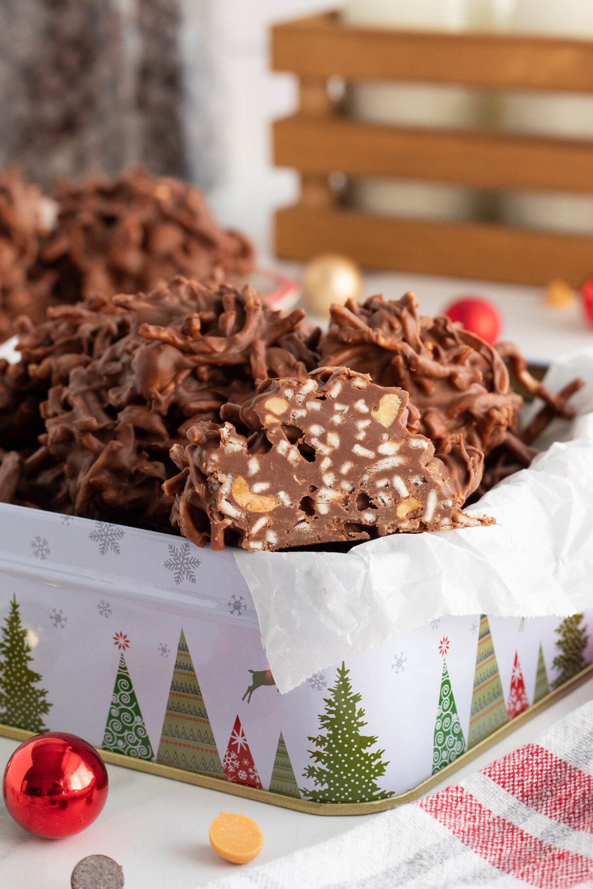 A festive tin lined with parchment paper is filled with clusters of chocolate-covered nuts. One cluster is cut open to show the nutty interior. Holiday decorations and ornaments are visible in the background.