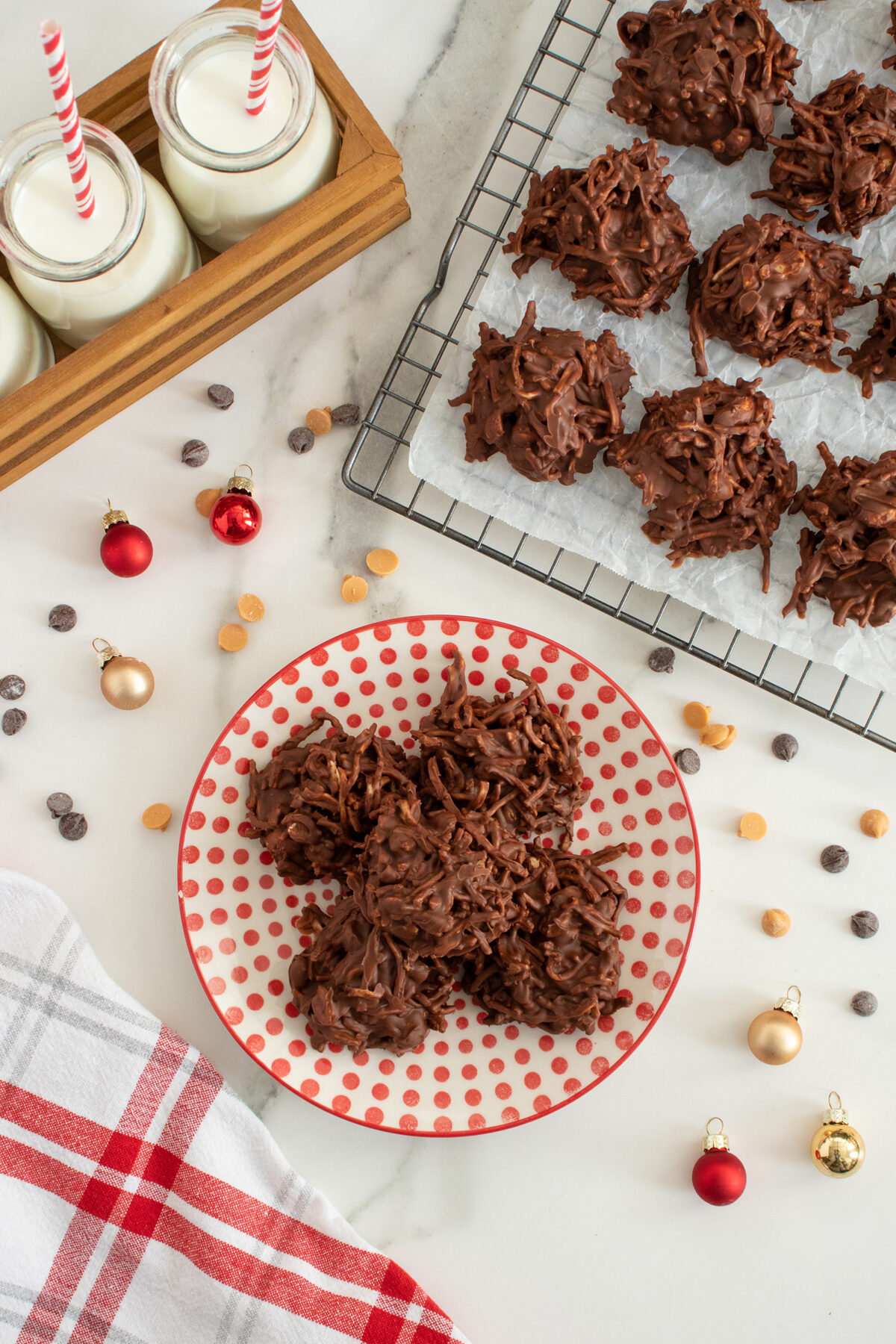 A red polka dot plate with chocolate haystack cookies on a white surface, next to a cooling rack with more cookies, glasses of milk with striped straws, and small Christmas ornaments scattered around.