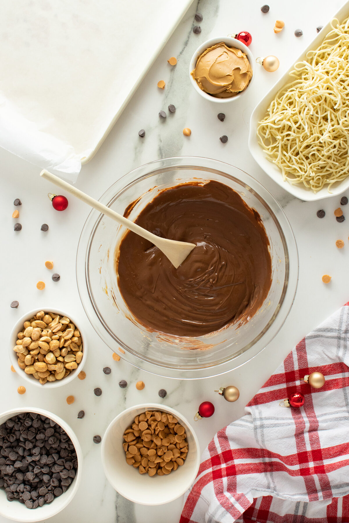 A glass bowl of melted chocolate with a spoon surrounded by bowls of chocolate chips, butterscotch chips, peanuts, peanut butter, and dry noodles on a white surface with a red plaid cloth and holiday ornaments.