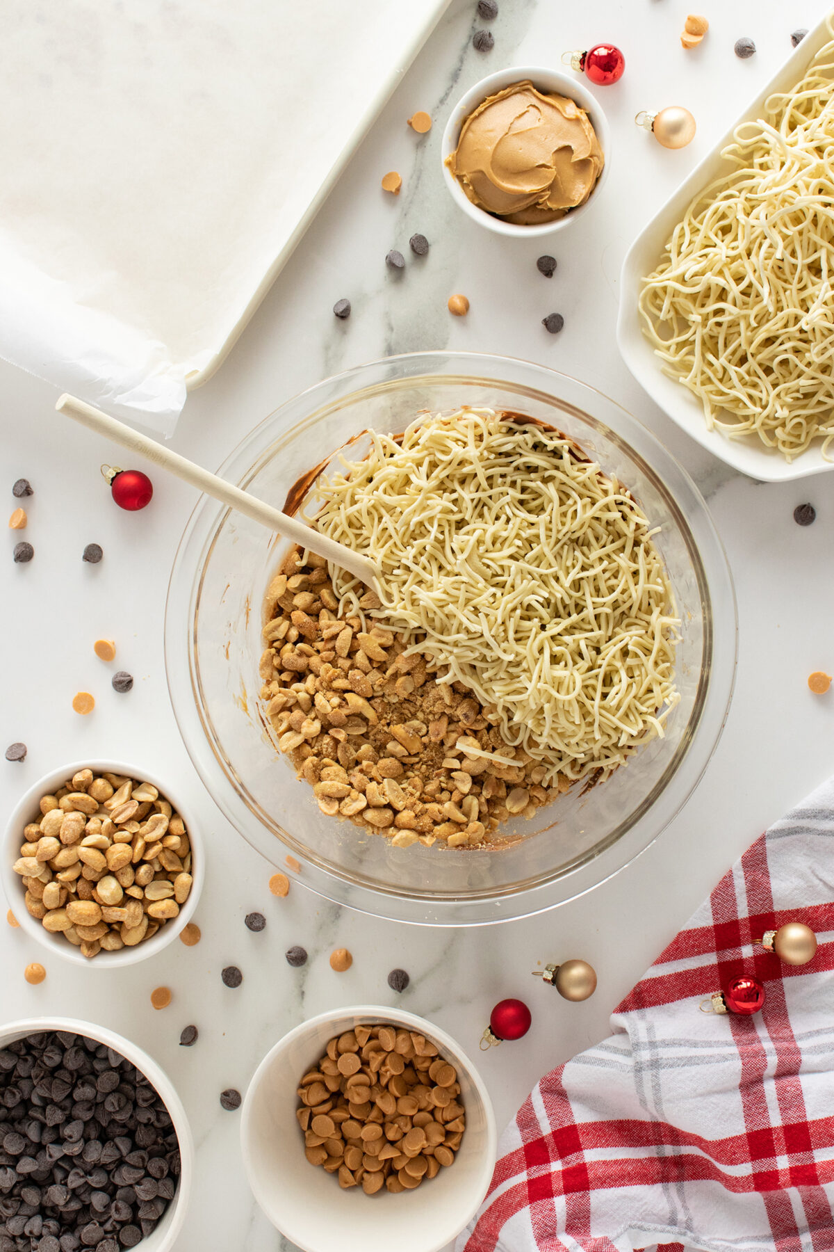 A glass bowl contains chow mein noodles, peanuts, and butterscotch chips being mixed. Surrounding the bowl are small bowls of peanuts, chocolate chips, butterscotch chips, and a plate of noodles on a white surface with festive ornaments.