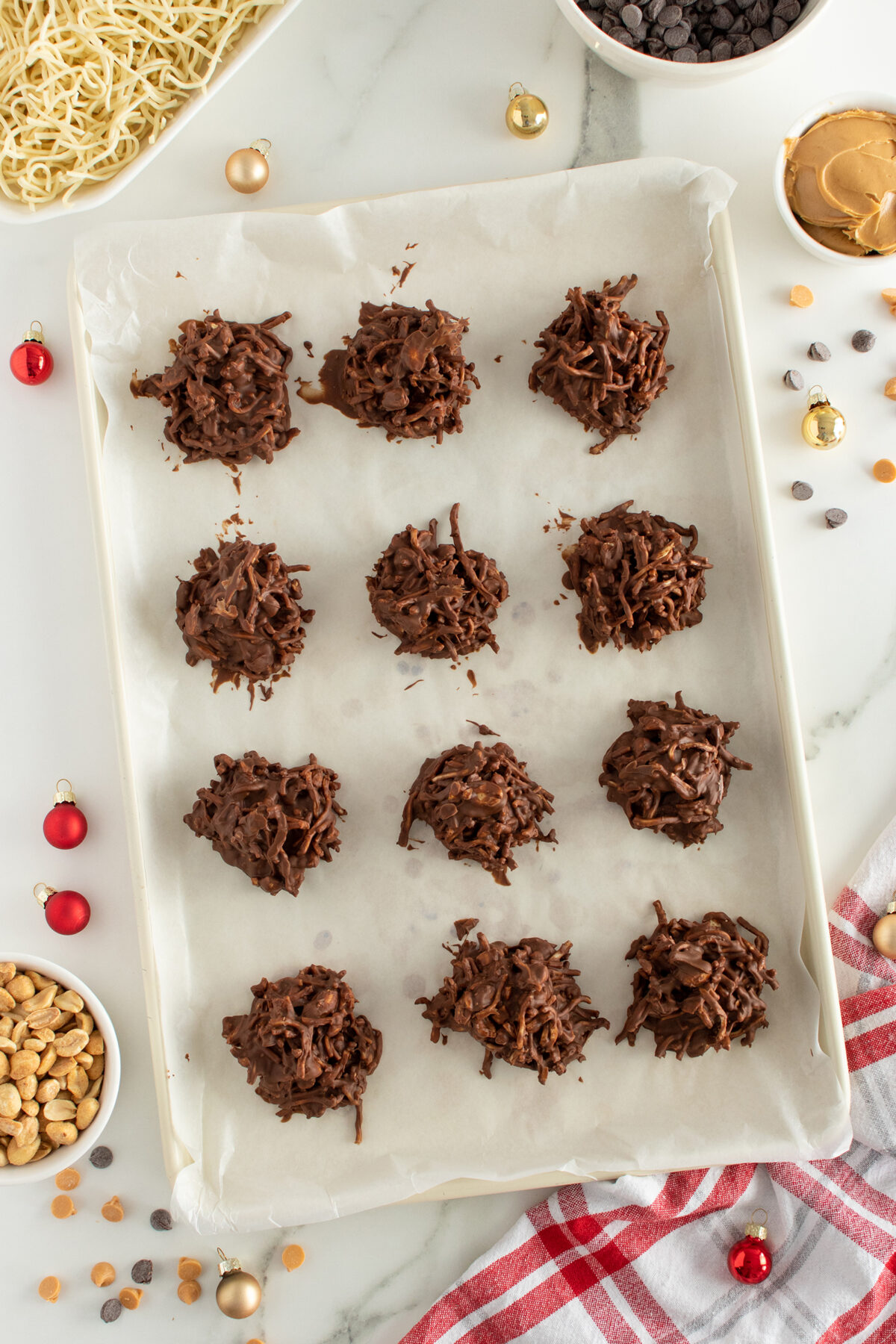 A baking sheet lined with parchment paper holds twelve chocolate haystack cookies. Surrounding the tray are bowls of peanuts, chocolate chips, peanut butter, uncooked noodles, and festive mini ornaments.