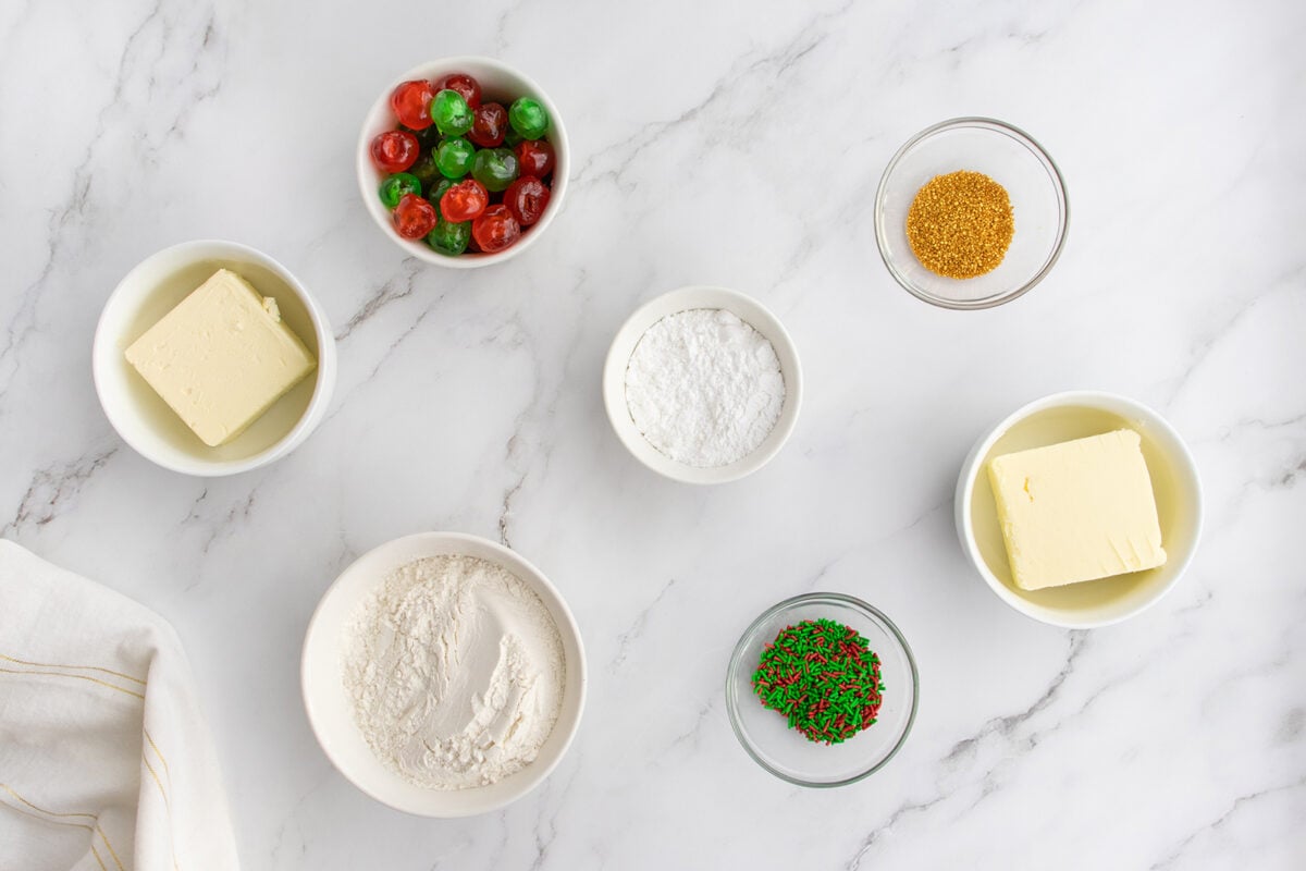 Seven small bowls on a marble surface contain butter, flour, powdered sugar, red and green candied cherries, gold sprinkles, and red-green sprinkles. A white cloth is visible in the bottom left corner.