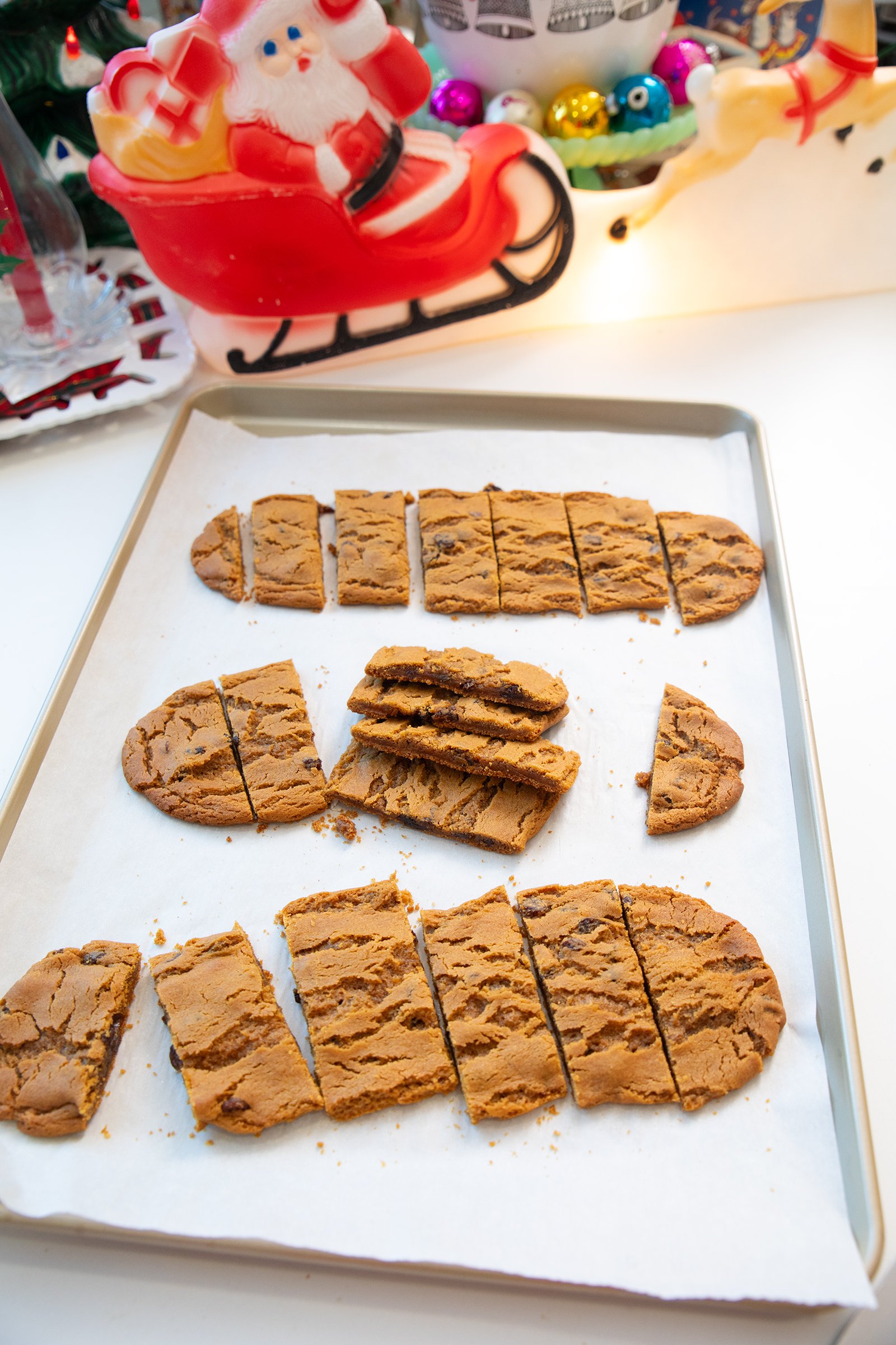 A baking tray with sliced, freshly baked biscotti cookies on parchment paper. In the background is a festive Santa Claus sleigh decoration and colorful Christmas ornaments.