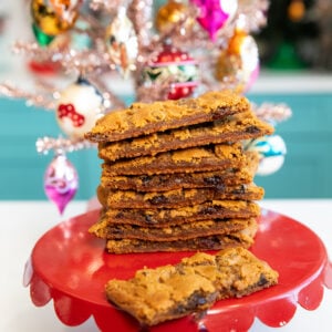 A stack of chocolate chip cookie bars sits on a red cake stand in front of a decorated, pink Christmas tree with colorful ornaments. One cookie bar is lying flat at the front of the stack.