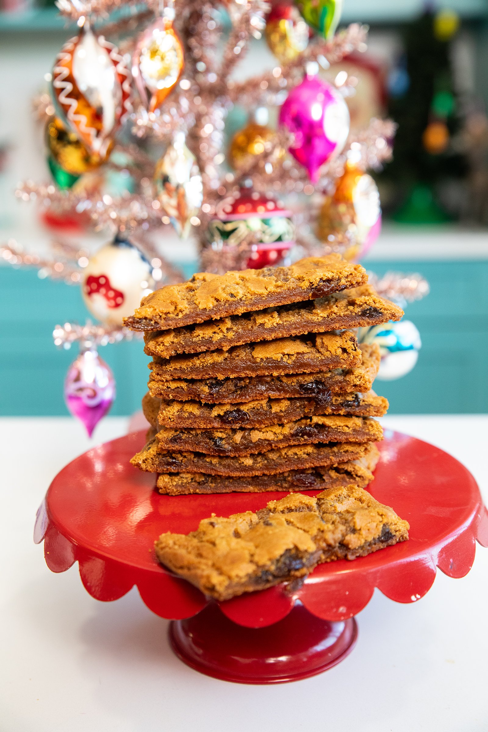 A stack of chocolate chip cookie bars sits on a red cake stand in front of a decorated, pink Christmas tree with colorful ornaments. One cookie bar is lying flat at the front of the stack.