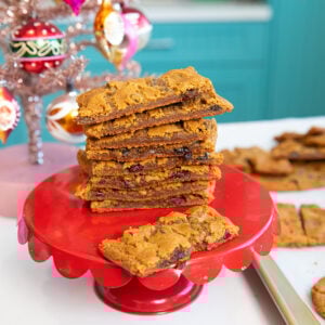 A stack of rectangular chocolate chip cookie bars sits on a red cake stand beside a silver Christmas tree with colorful ornaments; more cookies are on a baking tray in the background.