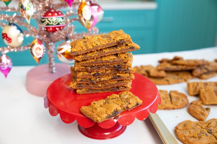 A stack of rectangular chocolate chip cookie bars sits on a red cake stand beside a silver Christmas tree with colorful ornaments; more cookies are on a baking tray in the background.