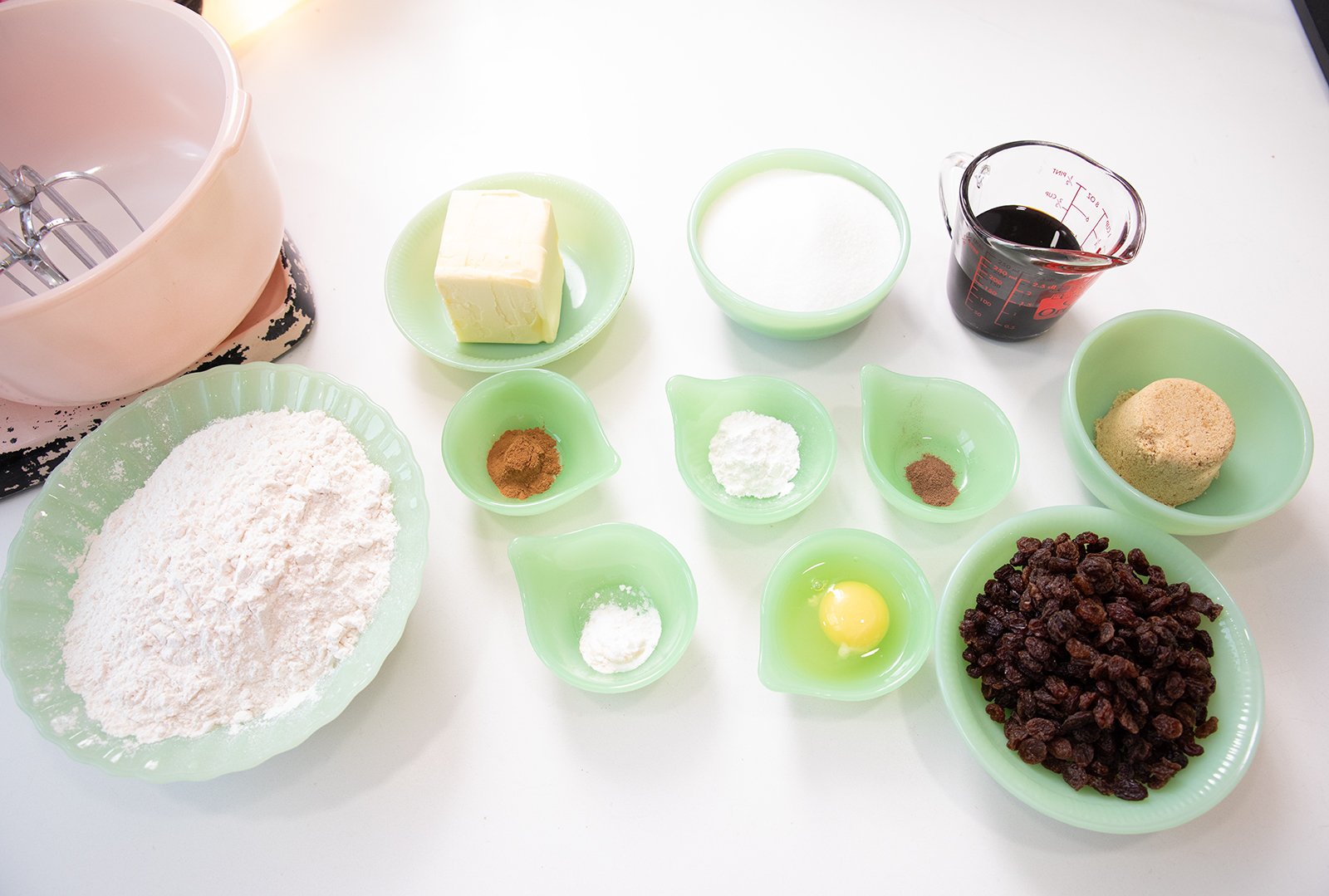 Baking ingredients on a white surface, including flour, butter, egg, raisins, brown and white sugar, cinnamon, nutmeg, baking powder, baking soda, vanilla extract, and a pink mixing bowl with a hand mixer.