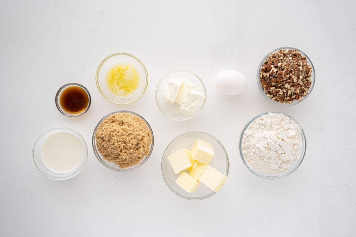 Various baking ingredients in glass bowls on a white surface, including butter, brown sugar, flour, chopped pecans, vanilla extract, an egg, milk, cream cheese, and melted butter.