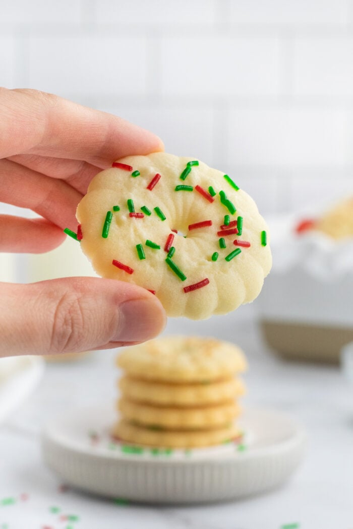 A hand holding a round, buttery cookie topped with red and green sprinkles. In the background, a stack of similar cookies sits on a white plate with a white tiled wall behind.