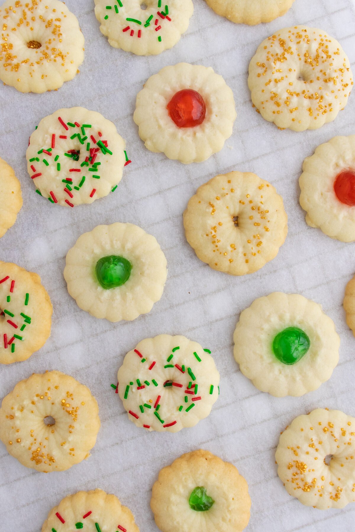 A variety of round spritz cookies on parchment paper, decorated with red and green sprinkles, yellow sugar, and red or green candied centers.