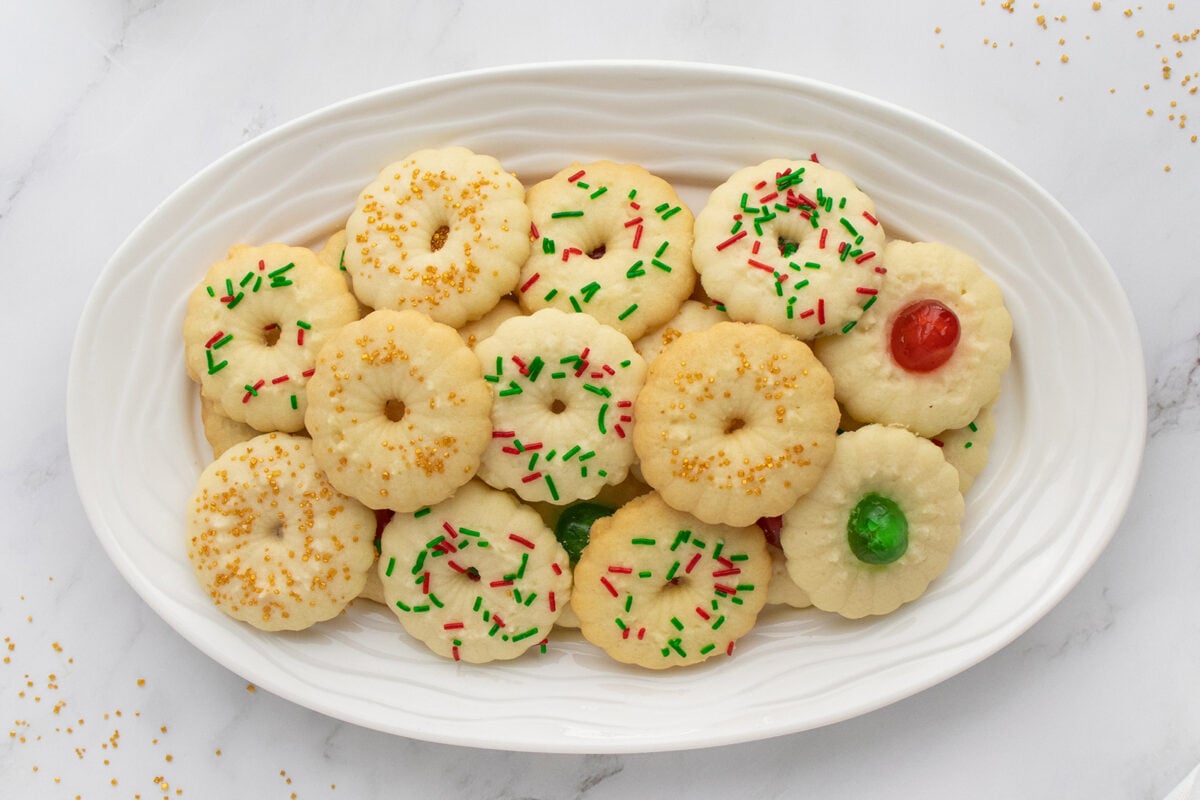 A white oval plate filled with round, festive spritz cookies decorated with red and green sprinkles, golden sugar, and red or green candied cherries on a white marble surface.