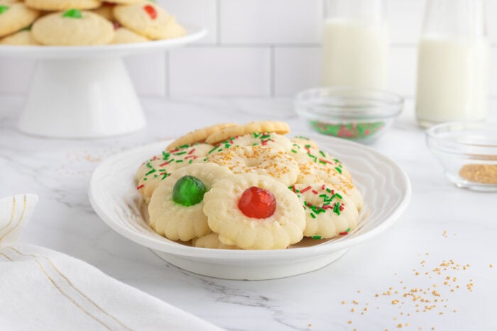 A plate of assorted spritz cookies topped with red and green candied cherries and colorful sprinkles, with more cookies in the background and bottles of milk on a marble countertop.