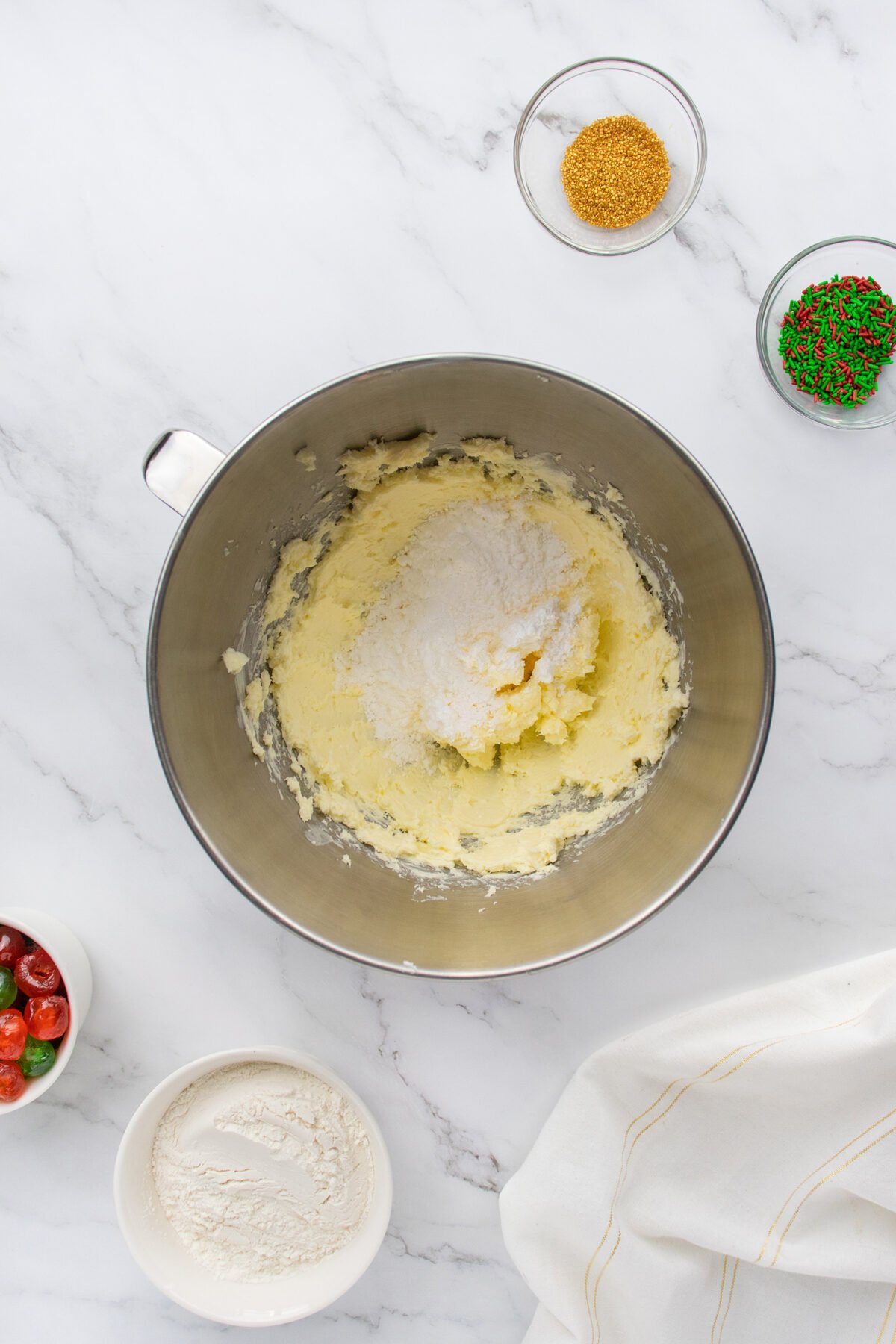 A mixing bowl with creamed butter and sugar sits on a marble countertop, surrounded by bowls of flour, red and green sprinkles, gold sugar, and maraschino cherries. A white towel is nearby.