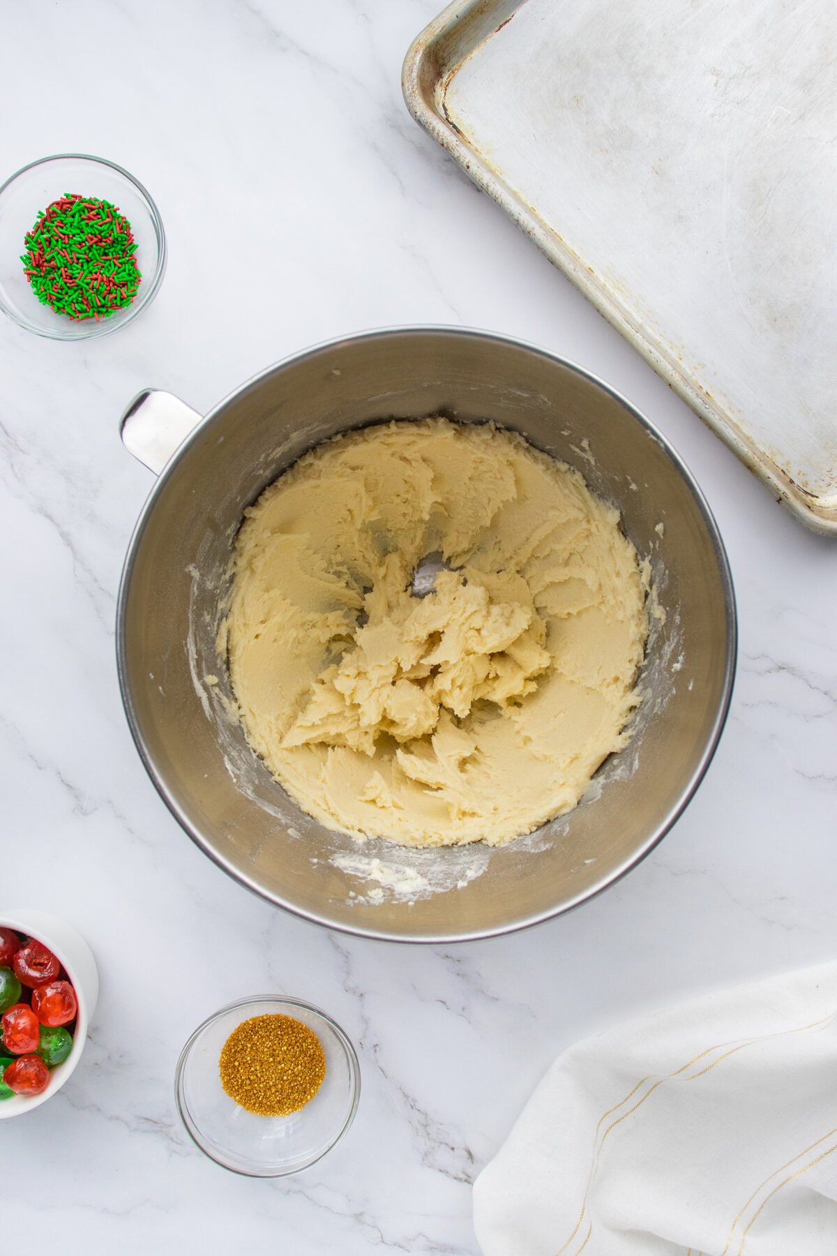 A metal mixing bowl with cookie dough sits on a marble countertop, surrounded by a baking sheet, a white towel, and small bowls of red and green sprinkles, gold sugar, and candied cherries.