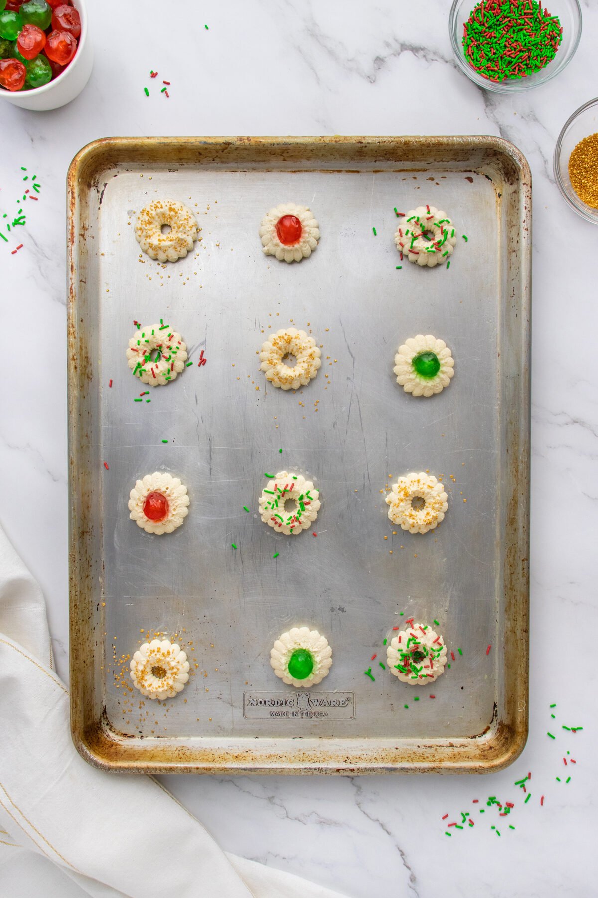 A baking sheet with twelve unbaked spritz cookies, each decorated with red or green candied cherries, gold sugar, or festive sprinkles, sits on a marble surface. Bowls of decorations are nearby, along with a white kitchen towel.